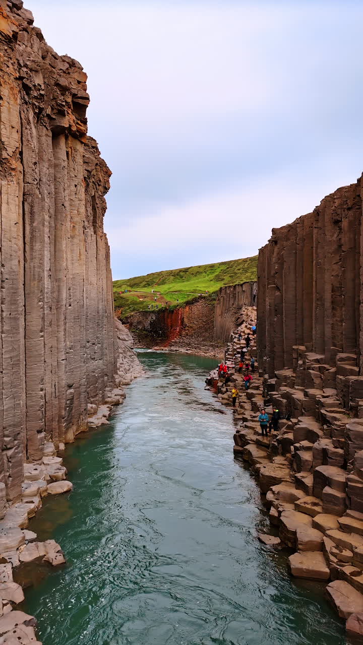 Tourists walk by the rocks near the narrow river. Beautiful columnar mountains in Iceland. Vertical video.