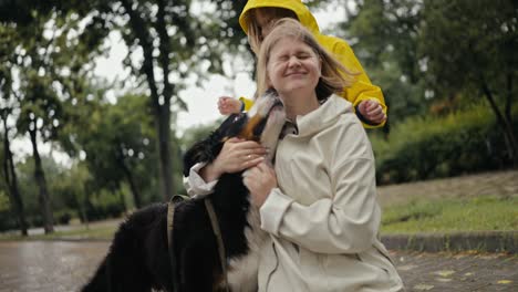 Happy-blonde-woman-in-a-white-jacket-plays-with-her-pet-black-dog-while-walking-with-her-daughter-in-a-yellow-jacket-along-the-alley-in-the-park-after-the-rain