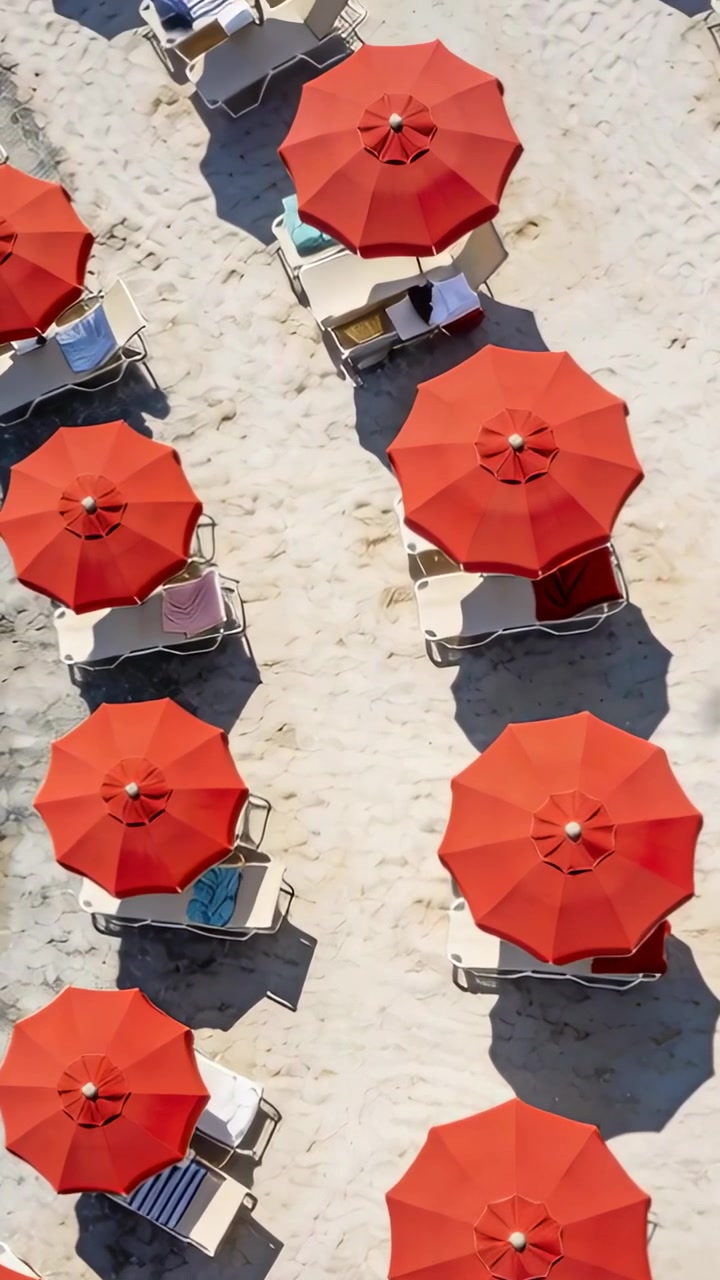 Video verticale: Drone che sorvola tende rosse sulla spiaggia, rivelando lettini bianchi.