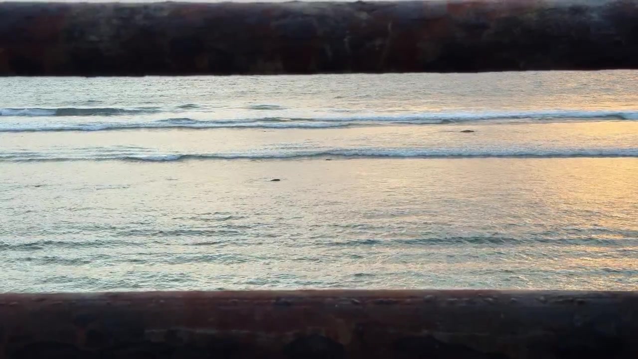 Onde oceaniche che si infrangono sulla riva al tramonto, viste attraverso una ringhiera