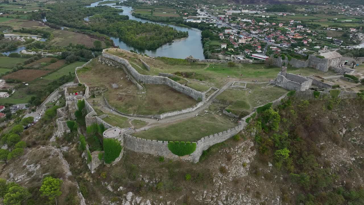 Aerial view of stone walls of Rozafa Castle atop a hill, overlooking winding river, green plains and city of Shkoder
