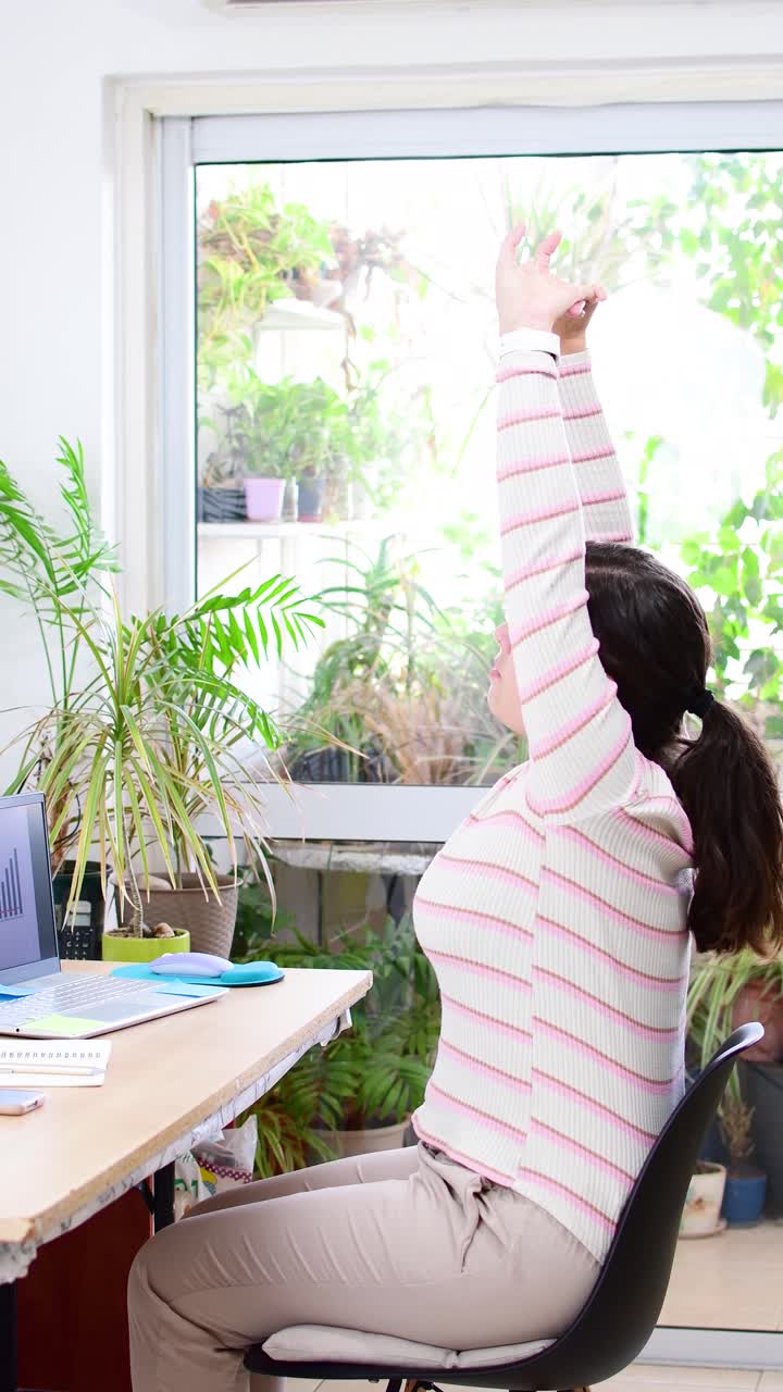 Waist up photo of young lady during a break sitting by the table and stretching her arms from hard work