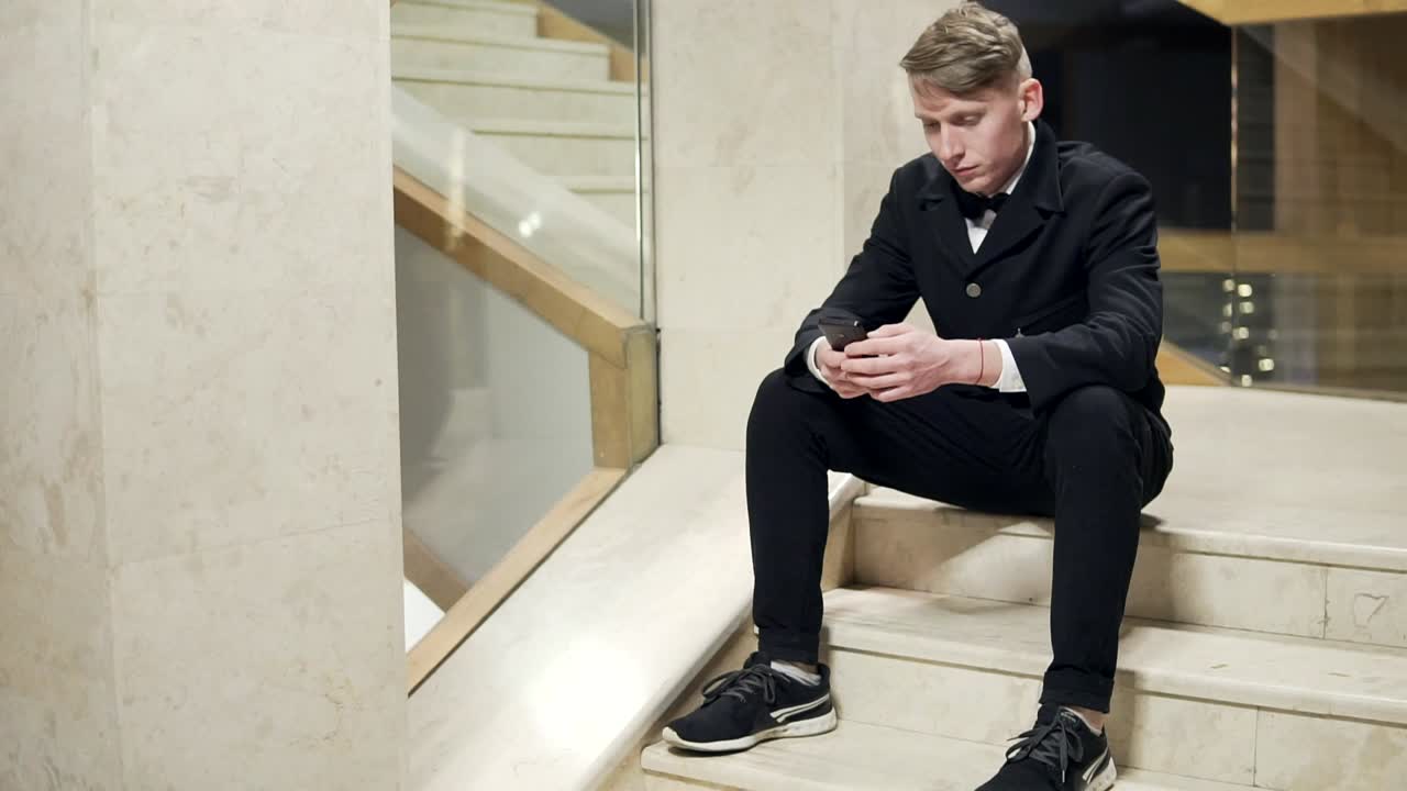 Young blonde man in black suit using his smartphone and sitting on stairs in hotel. Waiting for someone
