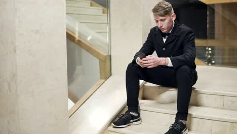 Young-blonde-man-in-black-suit-using-his-smartphone-and-sitting-on-stairs-in-hotel.-Waiting-for-someone