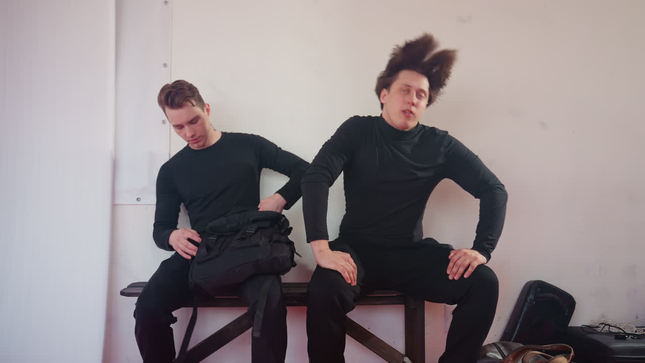 Two men inside room, one wearing backpack as they sit to change their outfit looking with serious expression, other with back turned in black shirt, plain background with white wall and hanging mat visible