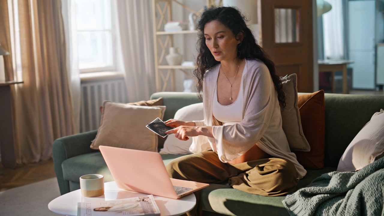 Futura mãe mostrando ultrassonografia em videochamada no laptop. Mulher grávida no sofá.