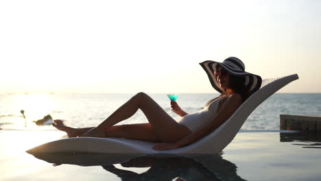 A-woman-relaxing-in-an-infinity-swimming-pool,-in-a-luxury-hotel,-enjoying-a-cocktail-at-sunset