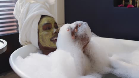 Smiling-african-american-woman-with-towel-and-mask-taking-bath-in-bathroom