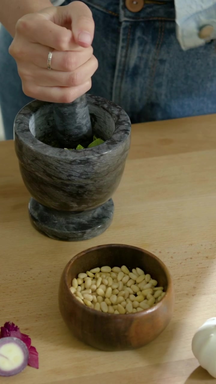 Vídeo vertical: Cozinheiro de camisa xadrez fatiando cebola, amassando ervas e cebola com gral e pilão para fazer pesto.