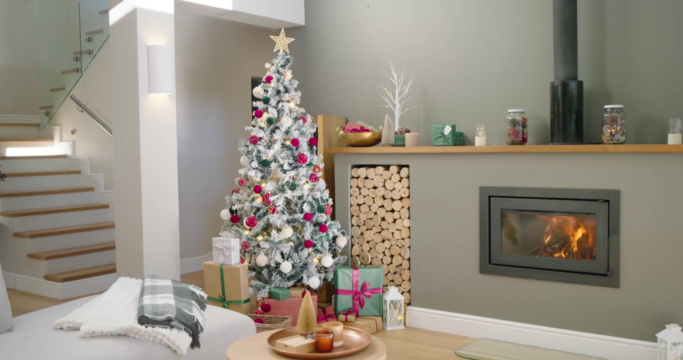 La telecamera esegue una panoramica e uno zoom su un albero di Natale innevato mentre il camino scoppietta, mostrando i regali
