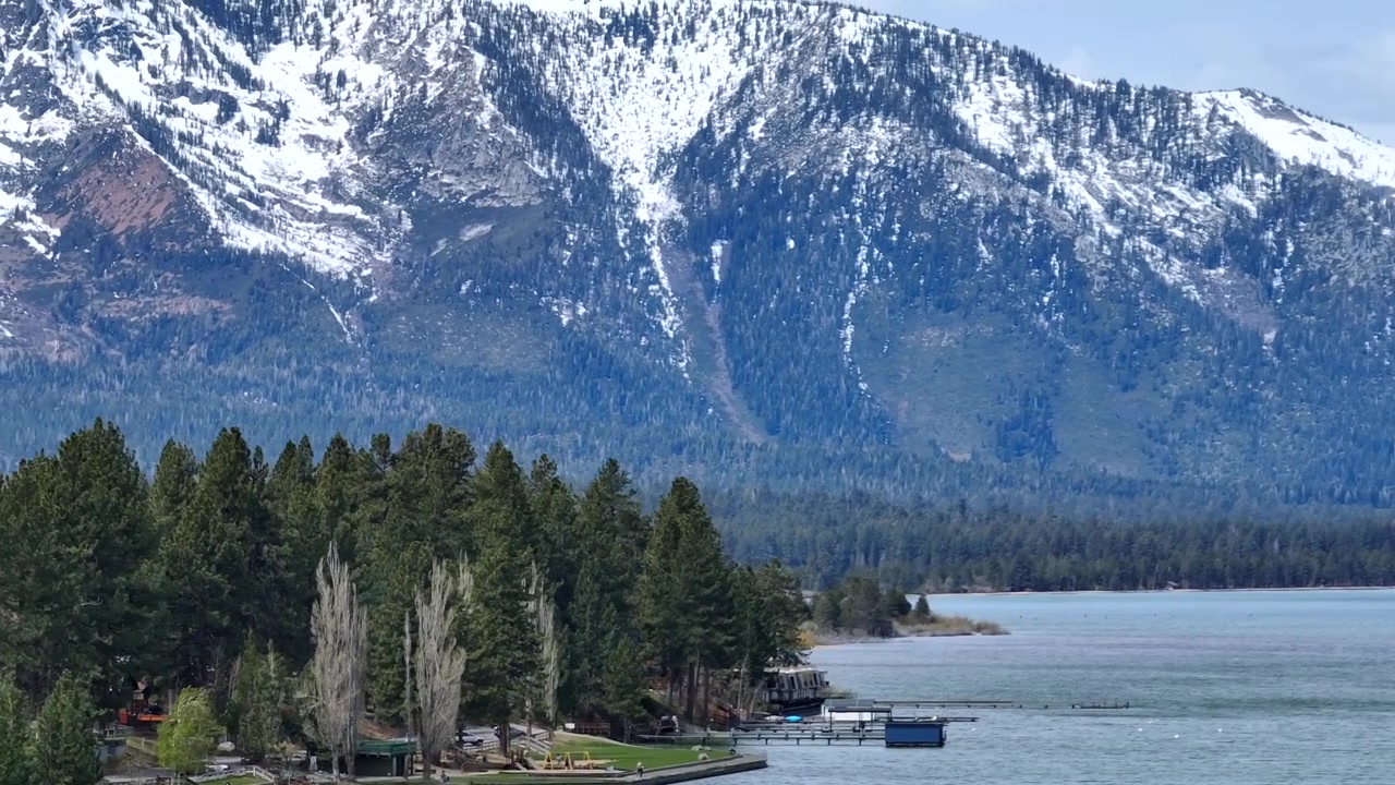 Panoramica costa del Lago Tahoe meridionale con pineta e acqua blu calma sotto le montagne innevate della Sierra Nevada in California, USA, pacifico paesaggio alpino ripreso dall'alto. Ampia ripresa aerea con zoom