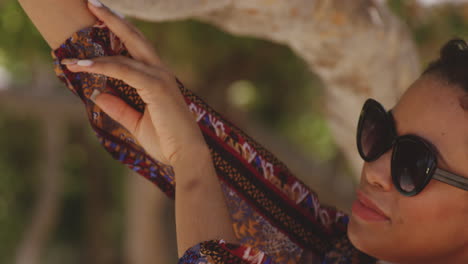 El-Primer-Plano-De-Una-Modelo-Latina-Con-Gafas-De-Sol-Posa-Junto-A-Un-árbol-Tropical-Junto-A-La-Playa,-Tiro-Revelador-Inclinado