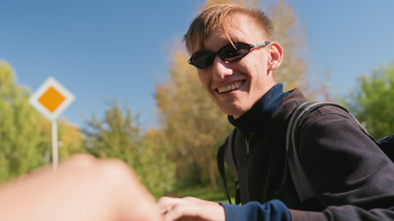 Kaukasische/r KurierIn lächelt und bietet eine High-Five auf einer sonnigen Vorortstraße in der Nähe eines Wegweisers an, spielerische Interaktion mit PassantIn, Austausch von Lachen und Gesten, trägt eine Sonnenbrille und einen Rucksack und liefert Essensbestellungen aus