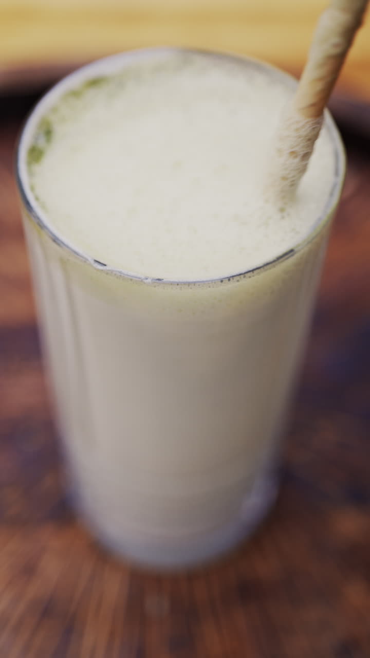 Close up of a woman mixing the foam on a matcha latte with a paper straw on a wooden tray. Vertical