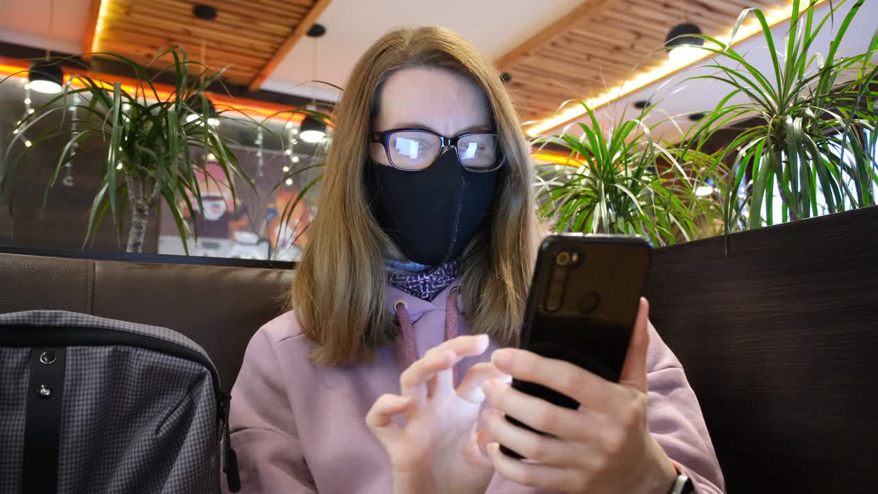 niña o mujer joven trabaja en un teléfono móvil en un café. mujer con una máscara protectora en la cara en una cafetería desplazándose a través de las redes sociales