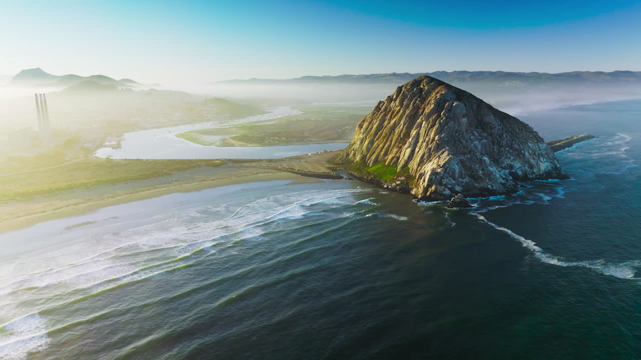 Flying closer to a pyramid-shaped mountain at the coast of Pacific. Little white waves coming slowly to the shore. White fog covering the horizon.