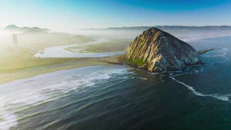 flying closer to a pyramid-shaped mountain at the coast of pacific. little white waves coming slowly to the shore. white fog covering the horizon.