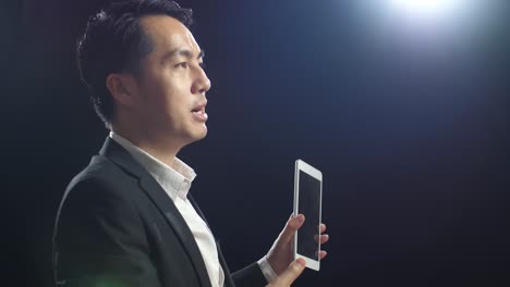 close up side view of asian speaker man in business suit holding and pointing tablet while speaking in the black screen studio