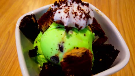 mint chocolate chip ice cream double scoop in a bowl surrounded by chocolate cake mini squares and topped with whipped cream and rotating on a wooden surface