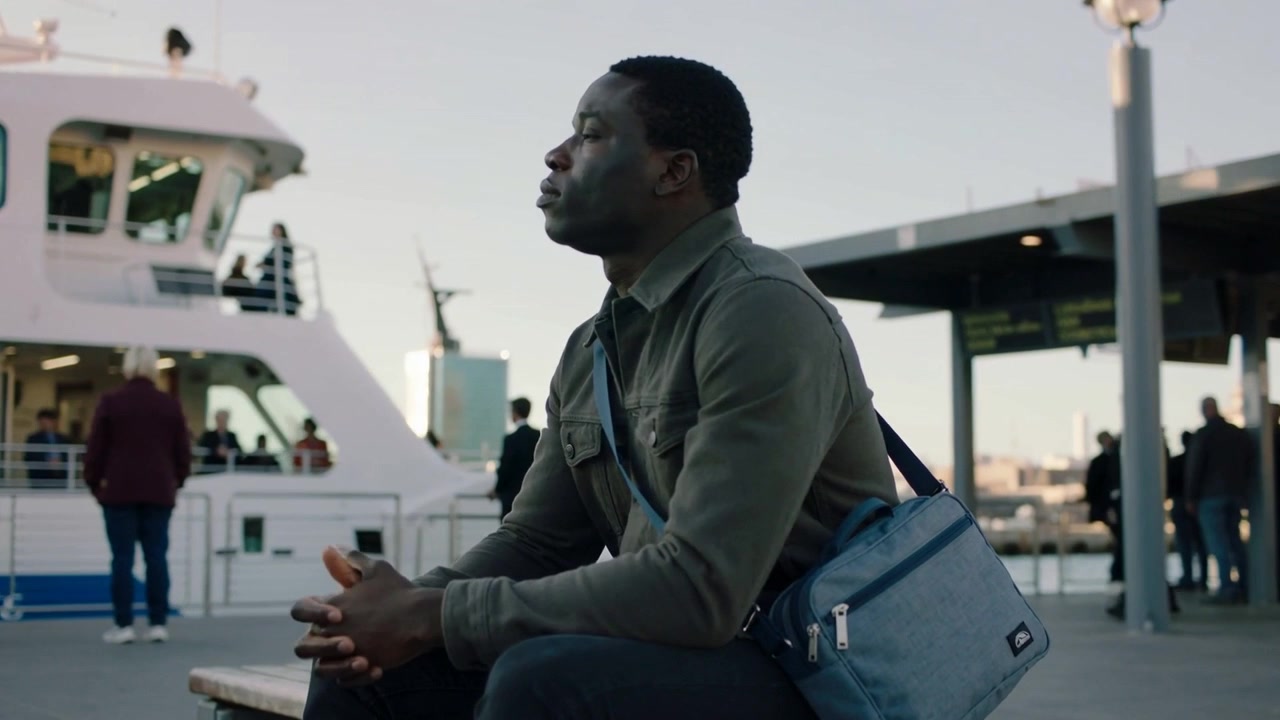 Sitting olive-jacket commuter adjusting blue crossbody strap on bench at ferry pier during boarding