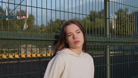 young woman leans on stadium fence to rest at sunset. sporty lady relaxes after jogging on sunny evening. active lifestyle and sadness from loneliness