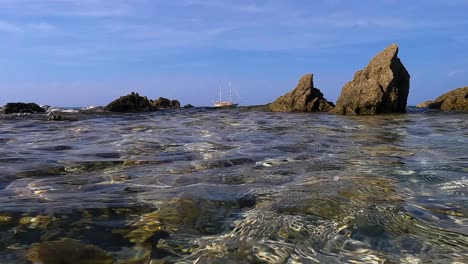 perspectiva de la superficie del agua de mar del velero amarrado en el horizonte con velas bajadas entre rocas