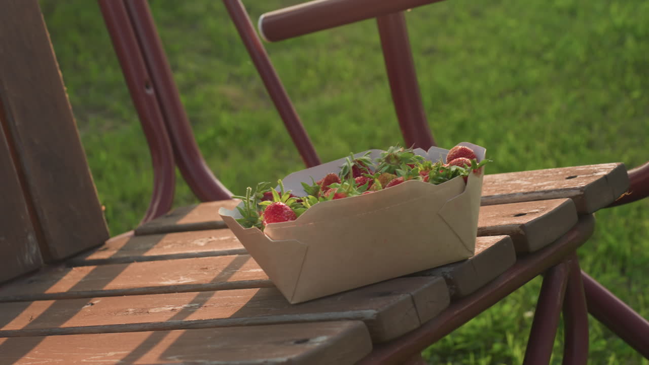 close up of fresh strawberries in cardboard container perched on wooden swing bench gently swaying above green grass during golden hour, warm sunlight casting soft glow on fruit and rustic bench