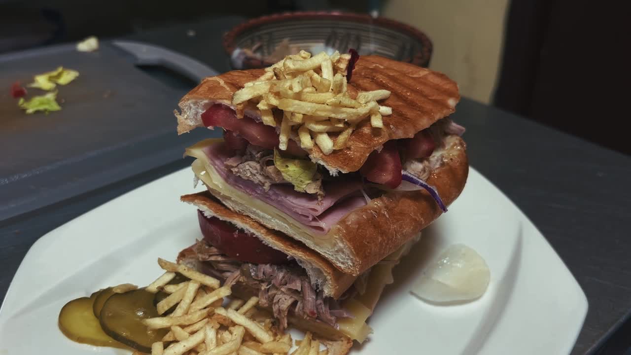 A large pressed sandwich sits on a plate, filled with layered ingredients and topped with thin fries, with pickles and scattered pieces visible on the counter