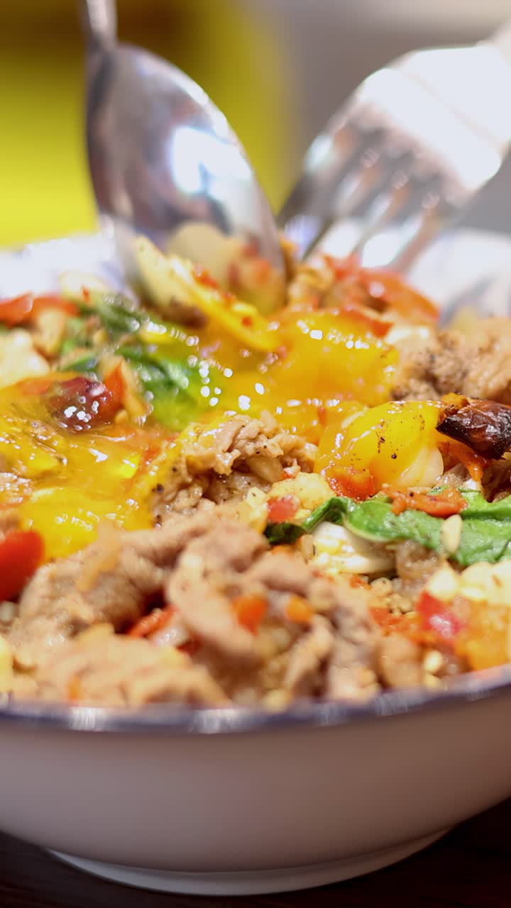 Primo piano di una forchetta e un cucchiaio che rompono un uovo con tuorlo liquido sopra una vivace ciotola di riso thailandese con gamberi, aglio, peperoncino ed erbe aromatiche sotto un'illuminazione calda.