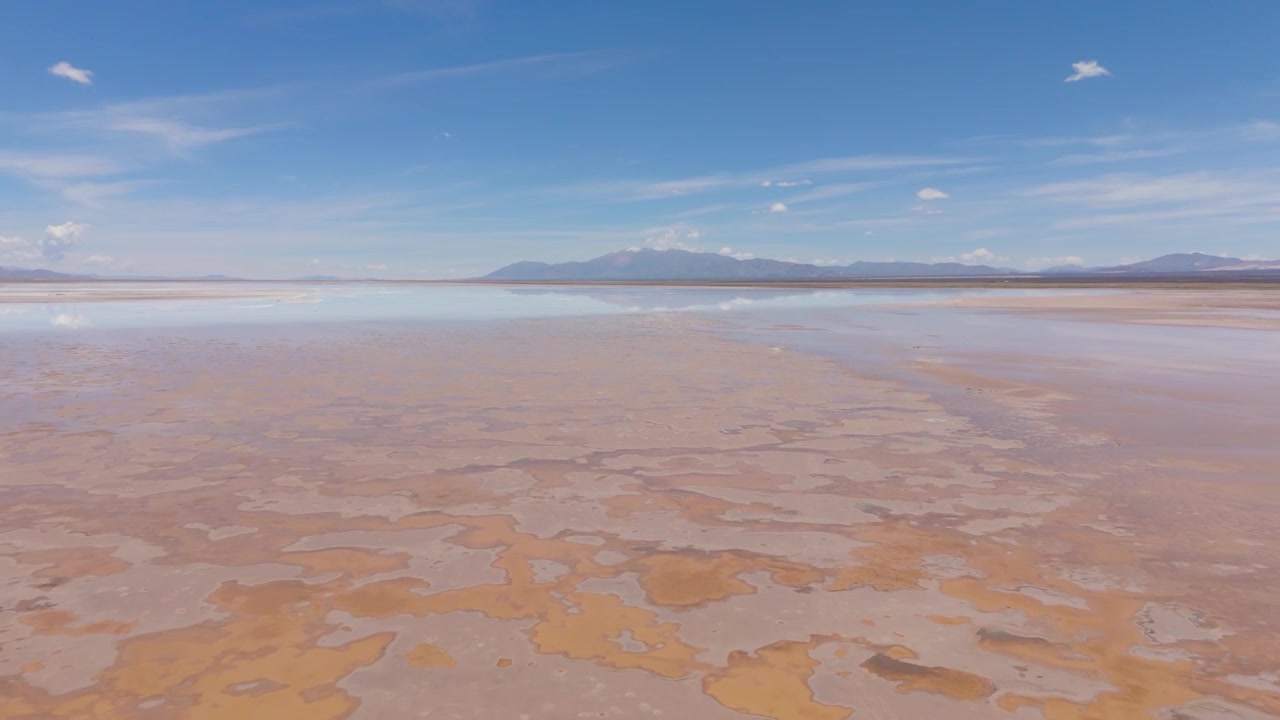Drone panorama above colorful Salinas Grandes salt flat with distant Andes, aerial upward tilt to ridges