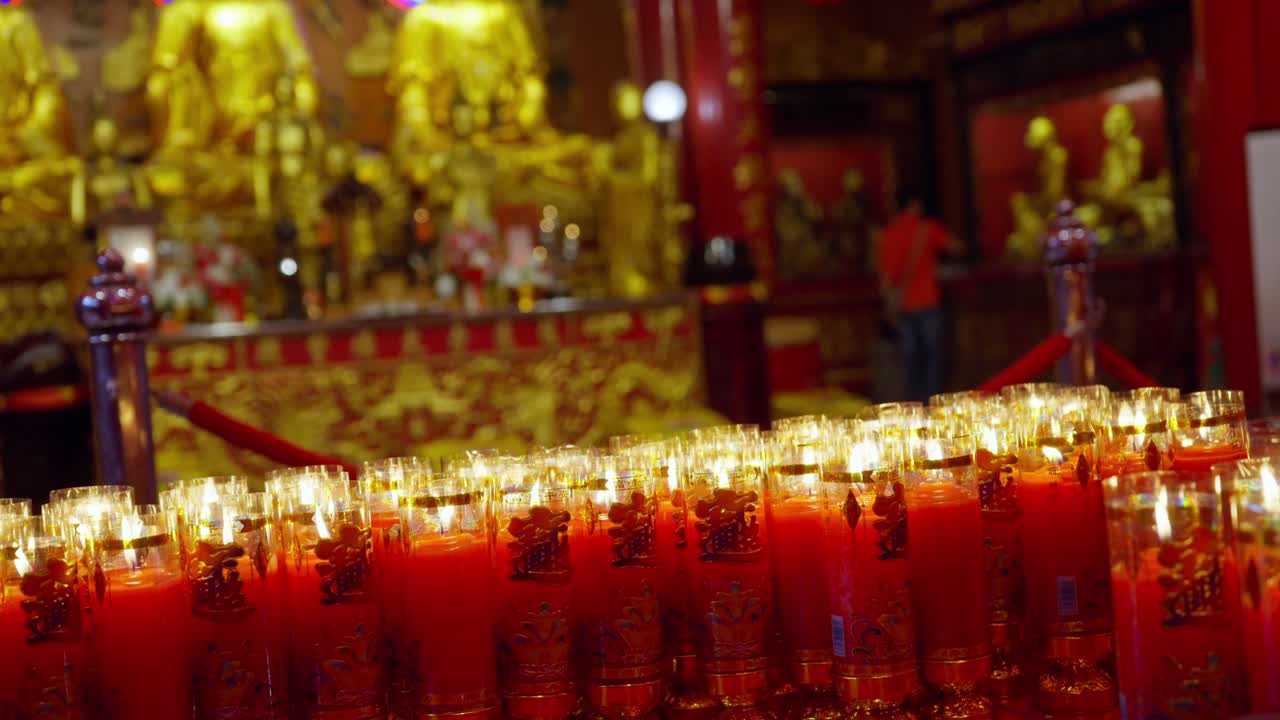Prayer candles lit and arranged inside ornate Chinatown temple, surrounded by golden decorations, slow motion flickering flames