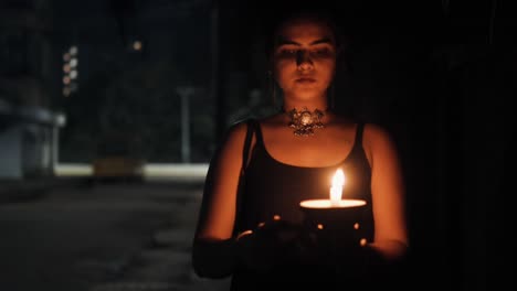 vista frontal de una mujer vestida de negro caminando por una calle oscura sosteniendo una vela encendida en las manos con el fondo borroso