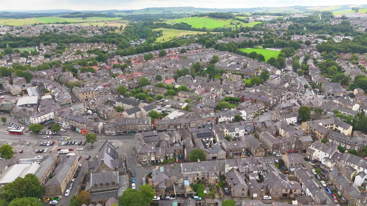 Riprese aeree scorrono fluidamente su una tradizionale città inglese con edifici in pietra, strade alberate e una campagna verdeggiante in lontananza sotto una luce diurna soffusa.