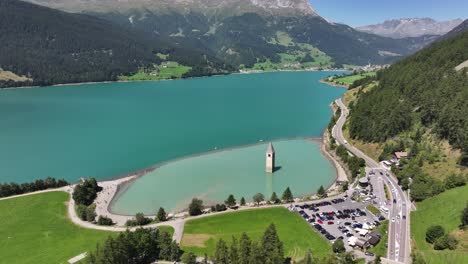 the famous bell tower in italy with a mountain backdrop and surrounding area, road, and vehicles, curon, italy