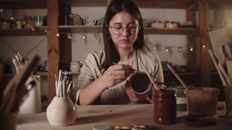 young woman potter drawing a design on the ceramic mug with a brush then looks in the camera