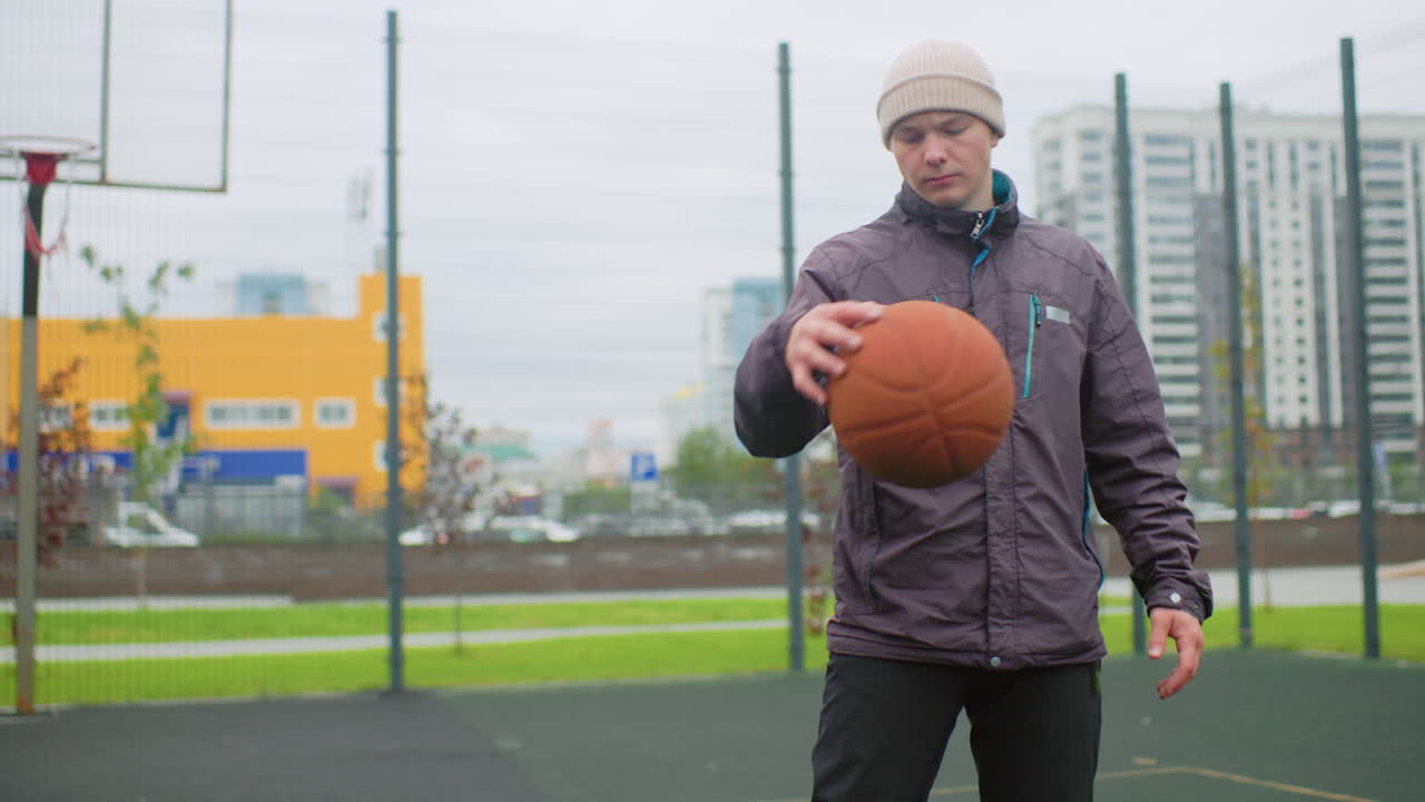Man dribbling basketball on court, overcast urban park with hoop and chainlink fence, wearing beanie and puffer jacket, solitary athlete practicing focused moves, residential towers in background