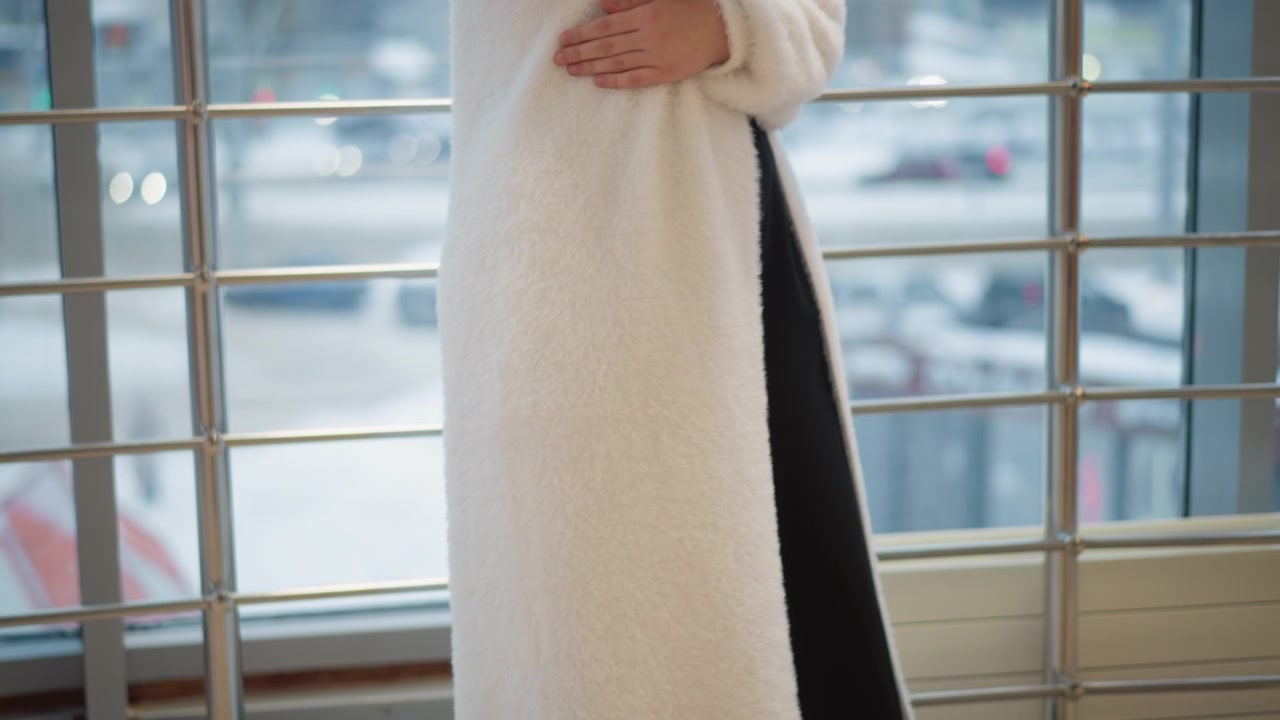 Vista dalla vita in giù di una persona che indossa un lungo cappotto bianco con pannello interno nero e orlo bordato di pelliccia, che si muove accanto a una ringhiera metallica e una finestra, con il passo, il sollevamento del tallone e il movimento fluido del capo.
