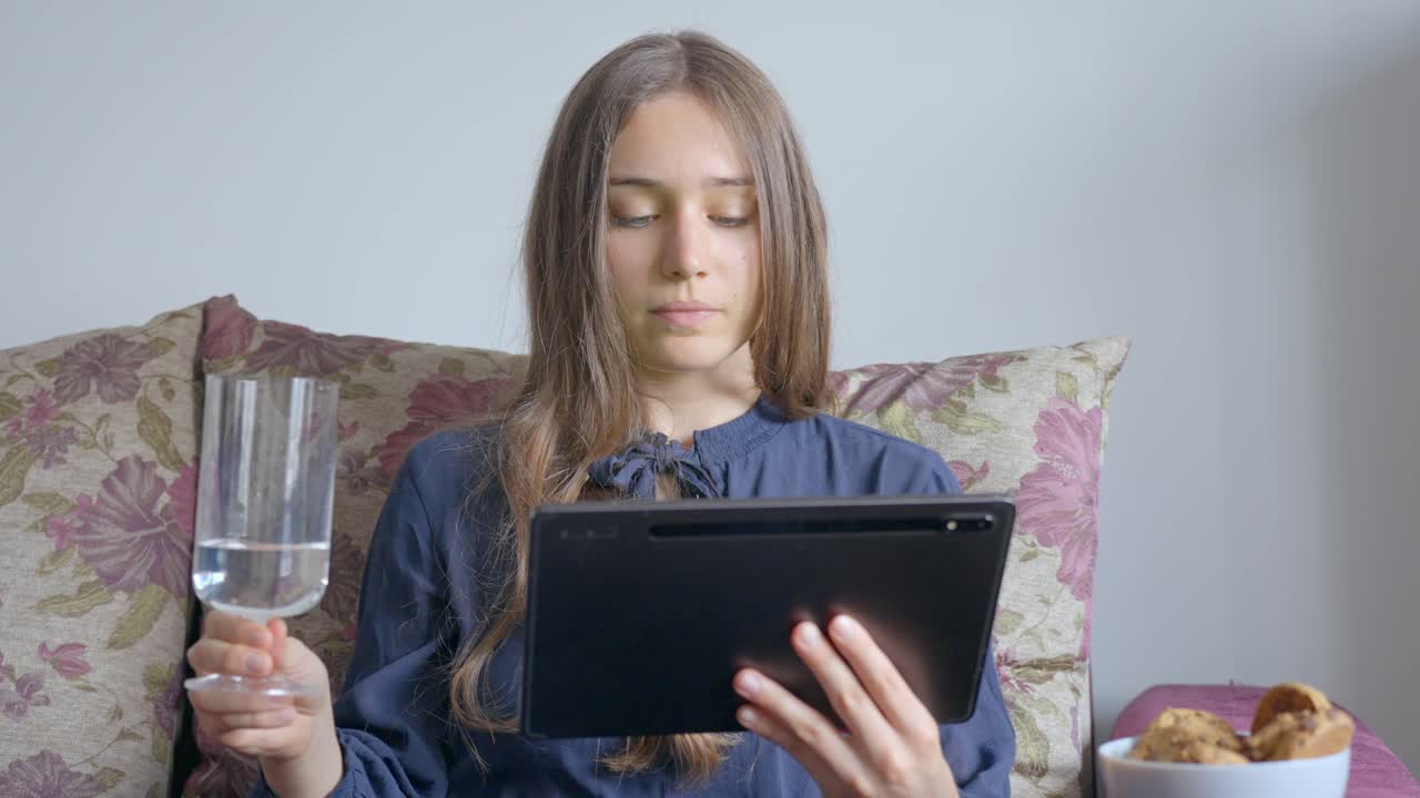 Young Woman sitting on the sofa with a glass of water and watching a tablet, in Slow Motion