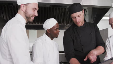 diverse male chef instructing group of trainee male chefs using tablet in kitchen, slow motion