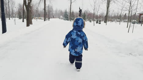 following the little kid wearing warm clothes walking by the winter park. toddler outdoors on snowing weather.