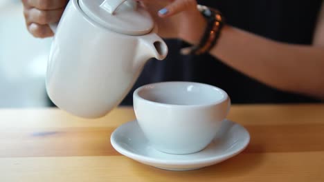 vista de cerca de las manos femeninas vertiendo té caliente de la tetera en la taza de porcelana blanca en la mesa de madera en la cafetería