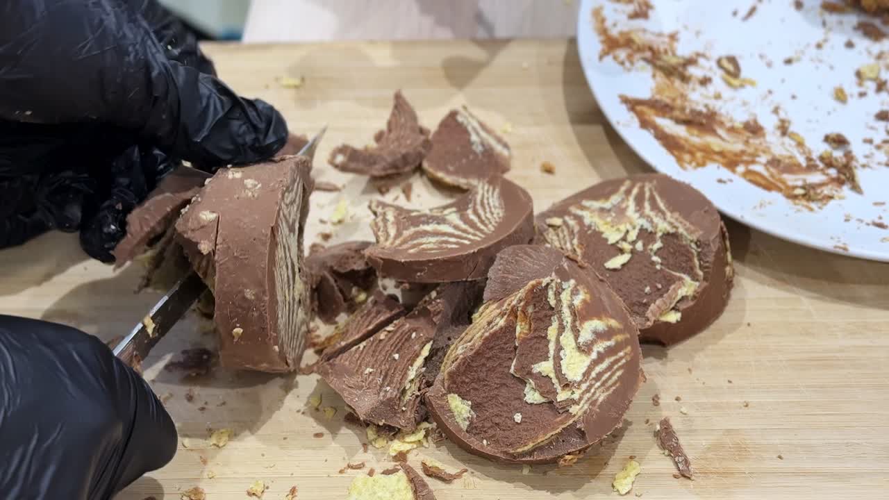 Primo piano di una mano guantata che taglia un dessert al cioccolato a strati su un tagliere di legno, con pezzi rotti, briciole, interno ripieno di crema e un piatto sporco in un ambiente di cucina interna