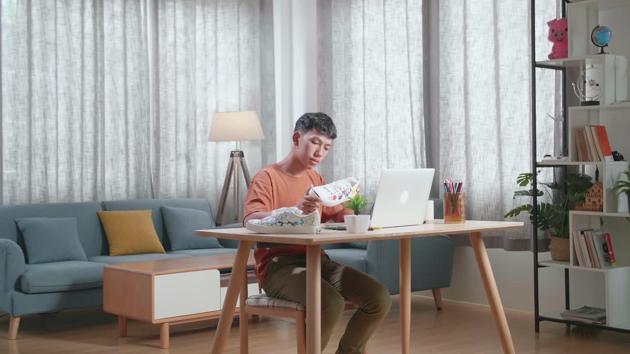 웃는 아시아 소년 신발 디자이너가 다채로운 패턴의 스니커를 들고 집에 있는 노트북의 사진과 비교합니다.