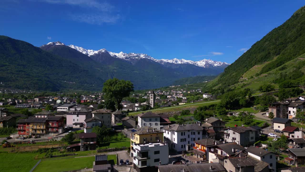 Colorful town beneath vibrant blue sky with snowy mountain peaks in the distance and a stone church in view. Shot in Villa di Tirano, Sondrio, Italy (Italia)