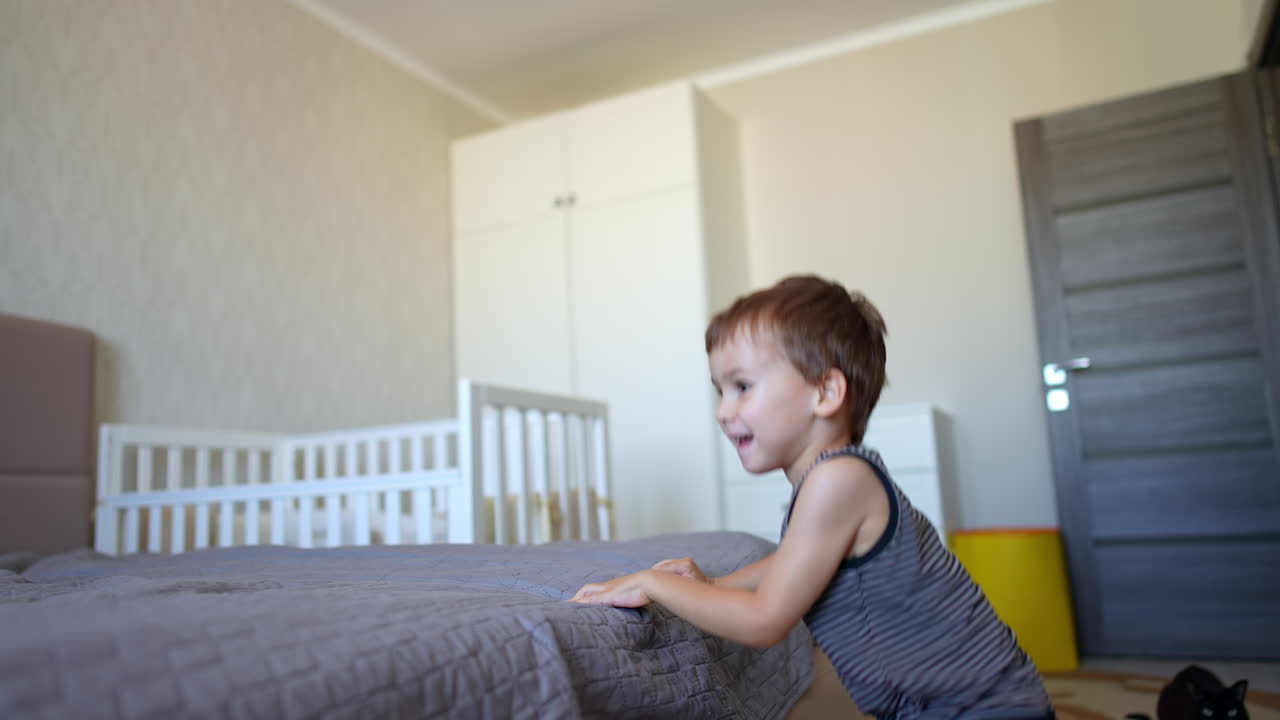Beautiful Caucasian toddler child climbs off the big bed. Baby boy in the bedroom. Black cat sits on the floor.