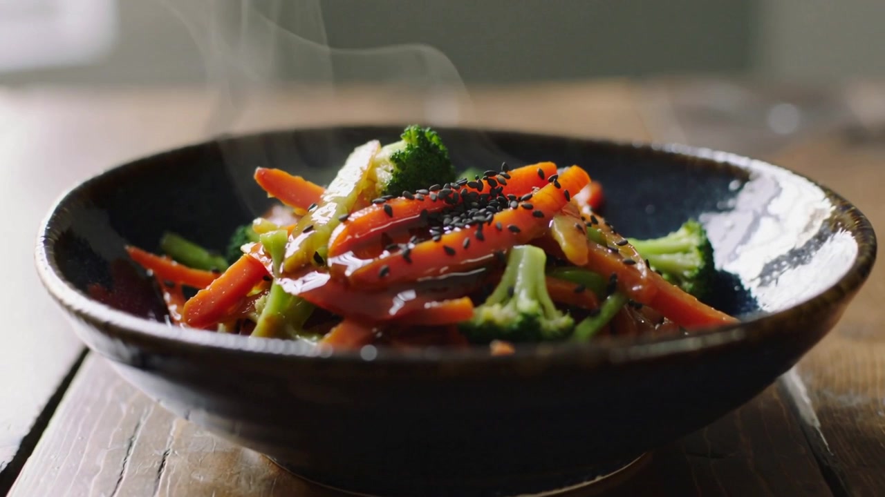 Telecamera che si allontana rivelando una ciotola fumante su un tavolo di legno, mostrando carote lucide, broccoli e sesamo.