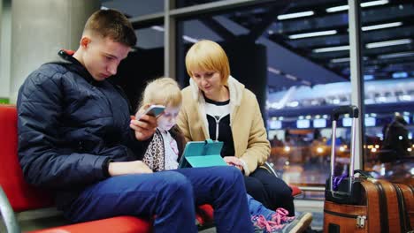 a family waiting for their flight at the airport terminal sitting in the waiting room using gadgets