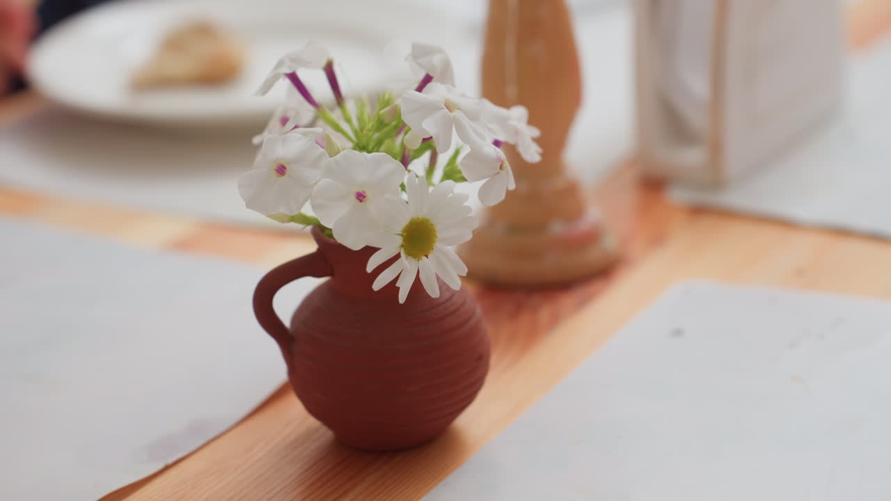 Un jarrón de arcilla con margaritas blancas y phlox sobre una mesa de aperitivos en una escuela infantil, un candelabro borroso al fondo, platos y servilleta visibles, la mano de un niño sostiene un pastel en el borde, una acogedora escena de aula, luz tenue.