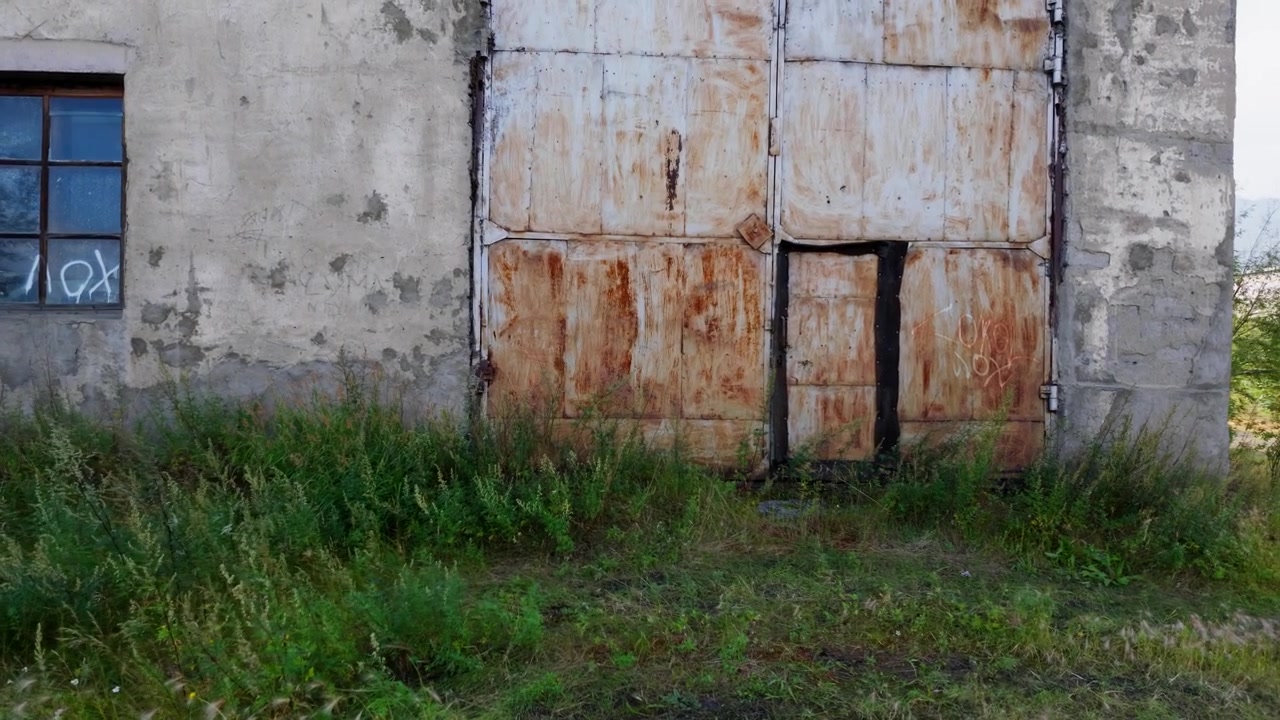 Una lenta panoramica verso destra rivela un edificio industriale arrugginito circondato da vegetazione incolta, con alberi e colline distanti visibili sotto un cielo luminoso.