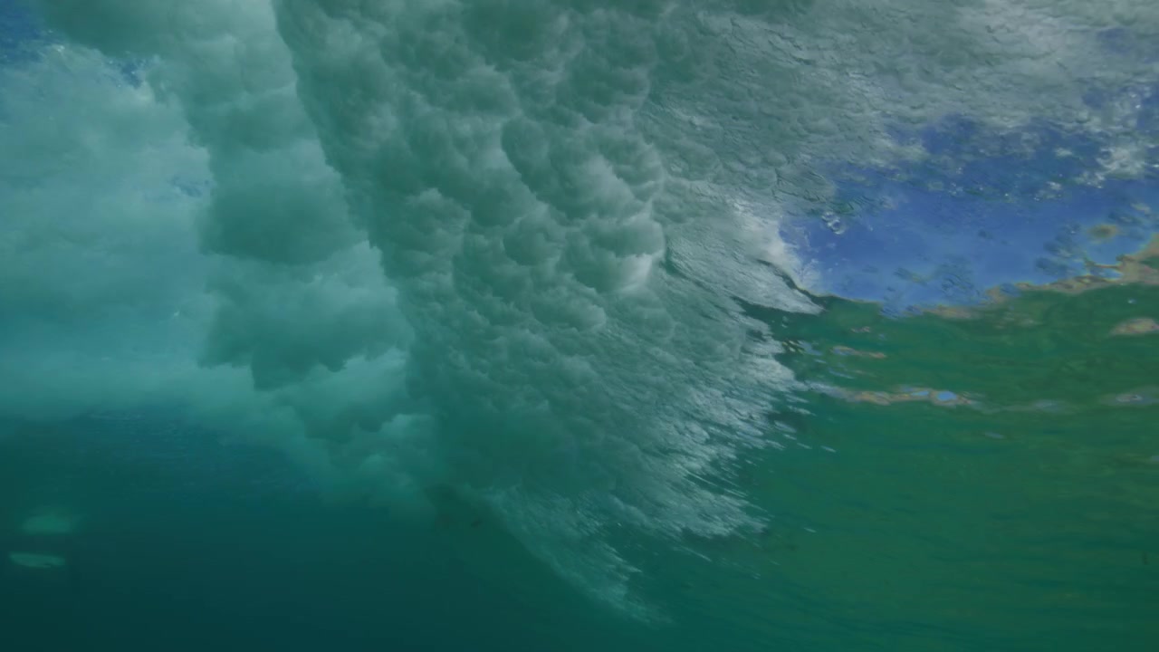Ocean wave rolls overhead through clear turquoise water with its curved surface seen from below, cloud whitewash vortex of bubbles spread